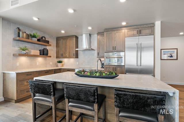 kitchen featuring wall chimney exhaust hood, a kitchen bar, built in refrigerator, a center island with sink, and decorative backsplash