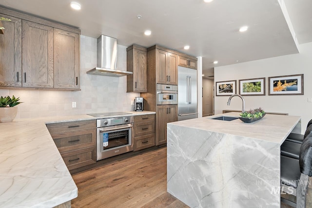 kitchen with sink, hardwood / wood-style flooring, appliances with stainless steel finishes, an island with sink, and wall chimney exhaust hood