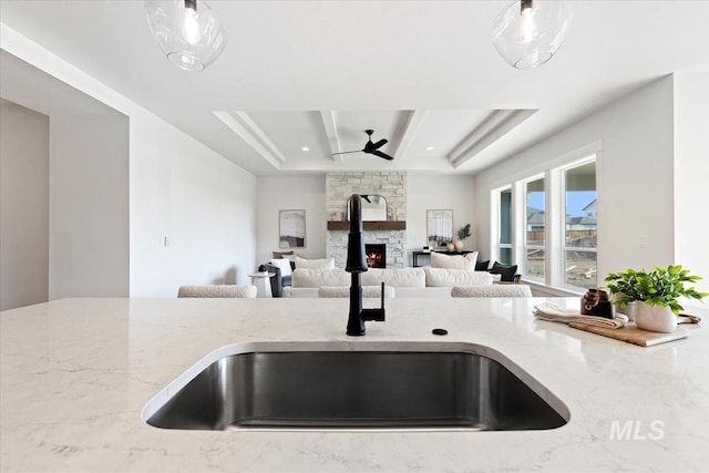 kitchen featuring a stone fireplace, sink, a raised ceiling, ceiling fan, and light stone countertops