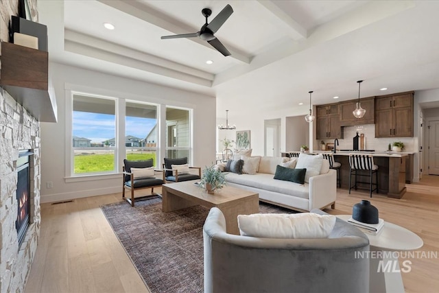 living room featuring a stone fireplace, ceiling fan with notable chandelier, beamed ceiling, a high ceiling, and light hardwood / wood-style floors