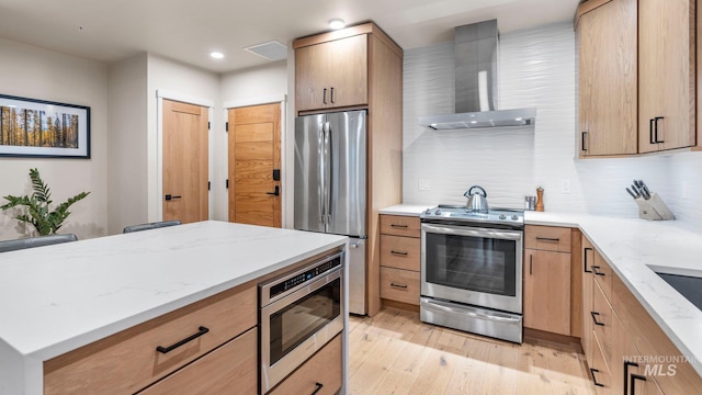 kitchen with light brown cabinets, light stone counters, light hardwood / wood-style floors, wall chimney exhaust hood, and stainless steel appliances