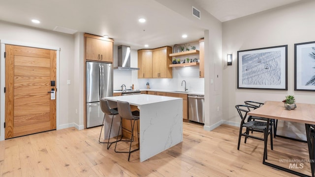 kitchen featuring wall chimney exhaust hood, a kitchen island, stainless steel appliances, and light wood-type flooring