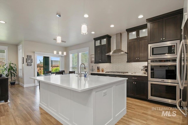 kitchen featuring wall chimney exhaust hood, an island with sink, decorative light fixtures, dark brown cabinetry, and stainless steel appliances