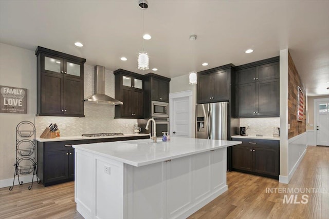 kitchen with sink, stainless steel appliances, wall chimney range hood, pendant lighting, and a center island with sink