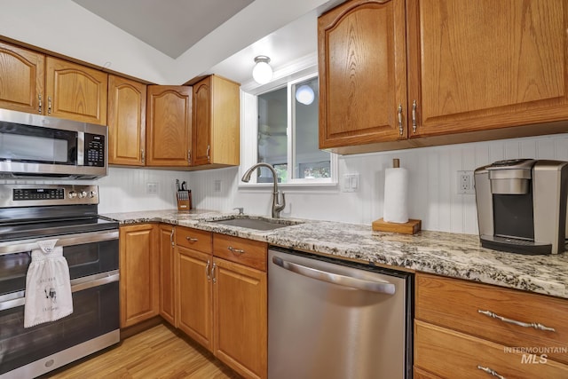 kitchen with appliances with stainless steel finishes, brown cabinetry, a sink, and light stone countertops