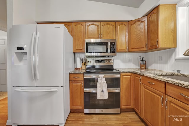 kitchen with stainless steel appliances, brown cabinetry, light wood-type flooring, and light stone countertops