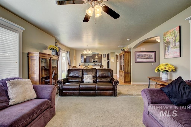 living room featuring baseboards, light colored carpet, ceiling fan with notable chandelier, arched walkways, and a textured ceiling