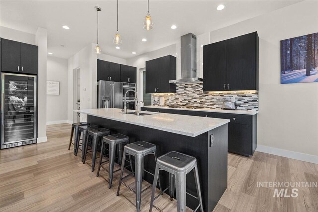 kitchen with hanging light fixtures, beverage cooler, wall chimney exhaust hood, a kitchen island with sink, and stainless steel appliances