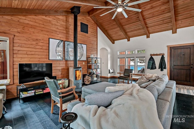 living room with ceiling fan, wooden walls, high vaulted ceiling, beamed ceiling, and a wood stove