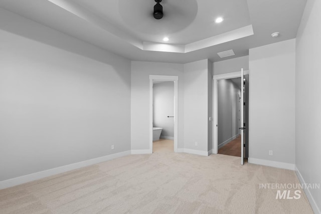 unfurnished bedroom with a raised ceiling, connected bathroom, light colored carpet, and ceiling fan