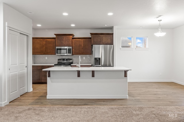 kitchen with light wood-type flooring, stainless steel appliances, sink, pendant lighting, and an island with sink