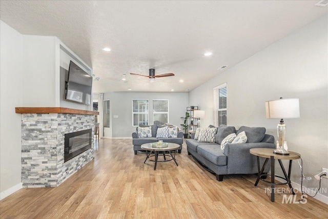 living area featuring a textured ceiling, a stone fireplace, light wood-style flooring, a ceiling fan, and baseboards