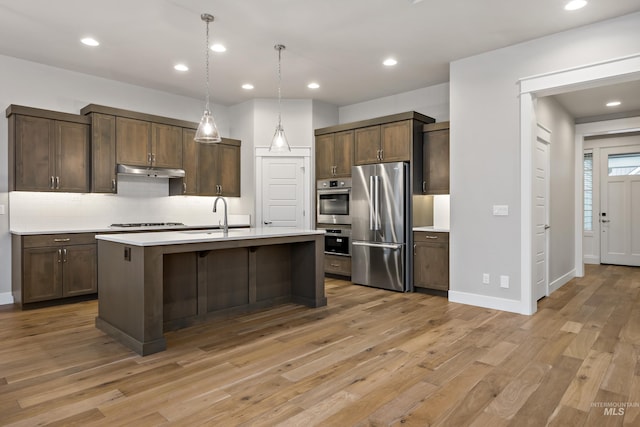 kitchen with appliances with stainless steel finishes, dark brown cabinets, pendant lighting, light hardwood / wood-style flooring, and an island with sink