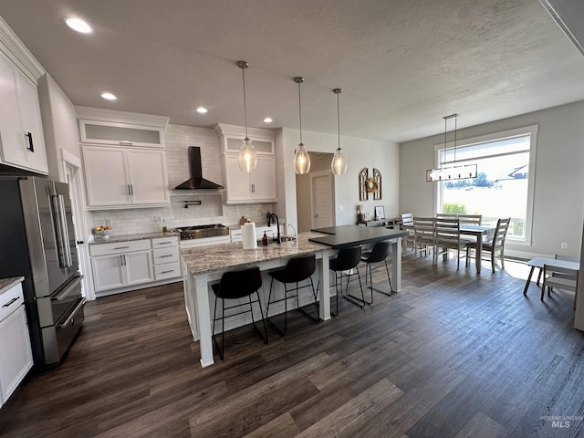 kitchen featuring wall chimney exhaust hood, stainless steel appliances, dark stone counters, a kitchen island with sink, and white cabinets