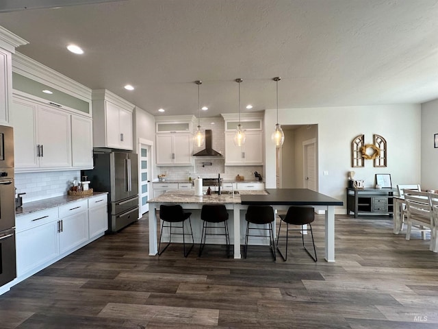 kitchen with white cabinetry, wall chimney exhaust hood, decorative light fixtures, high quality fridge, and a kitchen island with sink