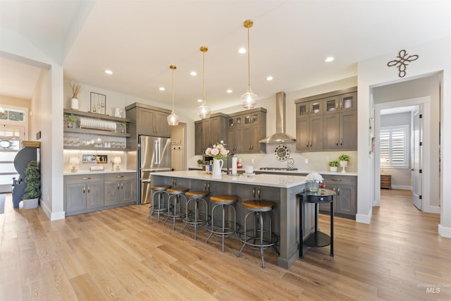 kitchen with stainless steel appliances, light countertops, wall chimney range hood, hanging light fixtures, and glass insert cabinets