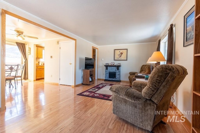 living area with a ceiling fan, baseboards, ornamental molding, a stone fireplace, and light wood-type flooring