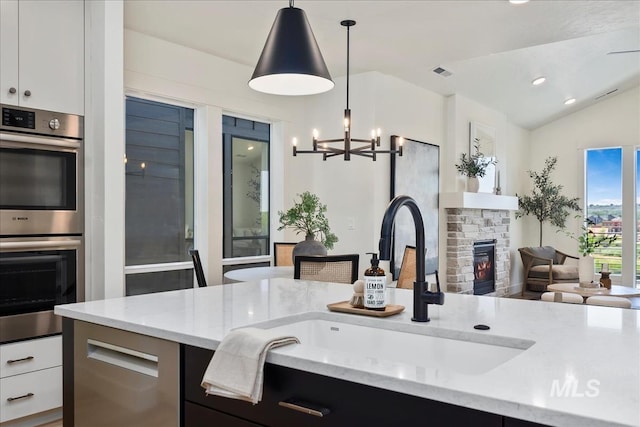 kitchen with light stone counters, double oven, white cabinets, a stone fireplace, and hanging light fixtures