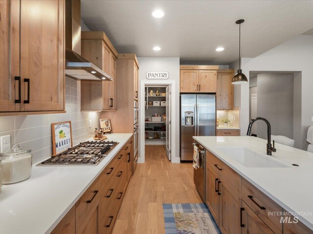 kitchen with a sink, stainless steel appliances, wall chimney range hood, light wood-type flooring, and light countertops