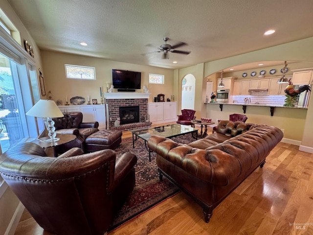 living area with a ceiling fan, a textured ceiling, arched walkways, a brick fireplace, and light wood-style floors
