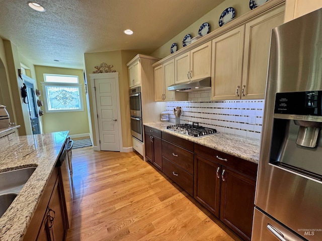 kitchen with under cabinet range hood, appliances with stainless steel finishes, decorative backsplash, and light wood-style flooring