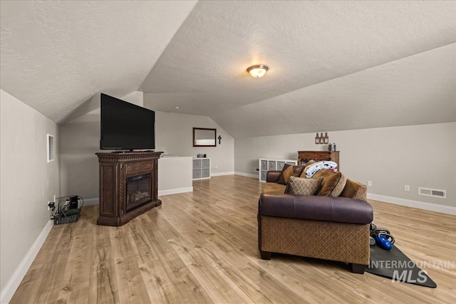 living room featuring a textured ceiling, light wood finished floors, vaulted ceiling, baseboards, and a glass covered fireplace