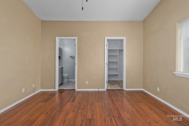 unfurnished bedroom with a closet, baseboards, ensuite bathroom, a walk in closet, and wood finished floors