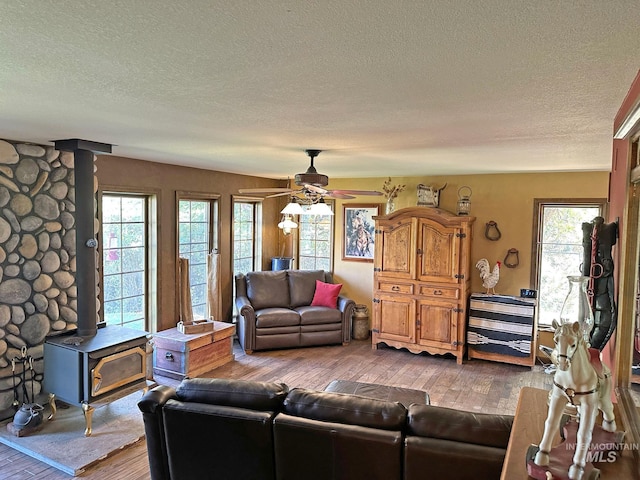 living room featuring a textured ceiling, a wood stove, wood-type flooring, a ceiling fan, and healthy amount of natural light