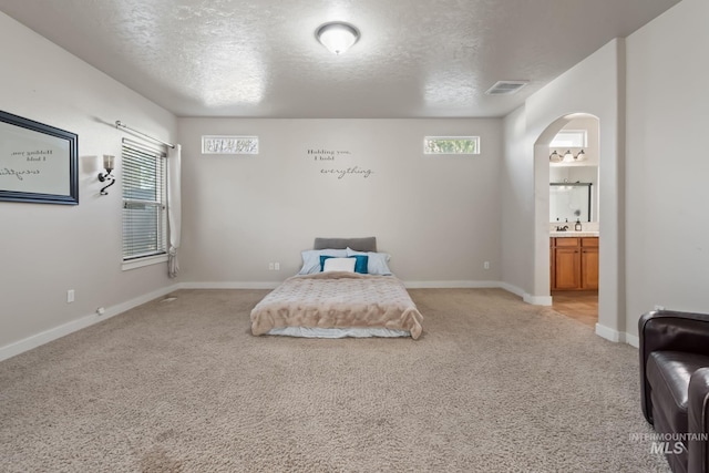 bedroom with connected bathroom, light colored carpet, arched walkways, a textured ceiling, and visible vents