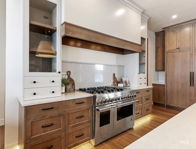 kitchen with custom exhaust hood, light wood-style flooring, glass insert cabinets, backsplash, and range with two ovens