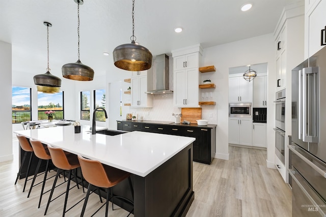 kitchen with wall chimney exhaust hood, open shelves, a sink, an island with sink, and recessed lighting