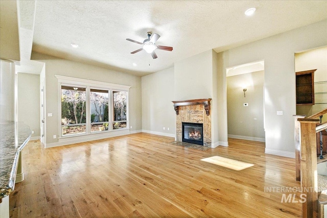 unfurnished living room featuring ceiling fan, a stone fireplace, a textured ceiling, and light hardwood / wood-style flooring