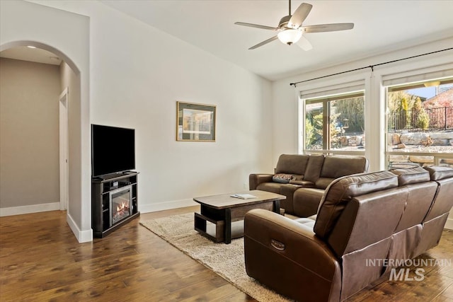 living area featuring a glass covered fireplace, wood finished floors, baseboards, and ceiling fan