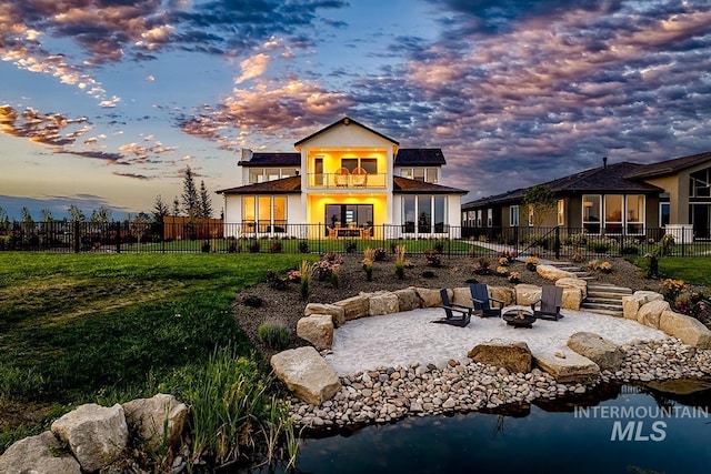 back house at dusk featuring a garden pond, a balcony, a yard, a patio area, and an outdoor fire pit
