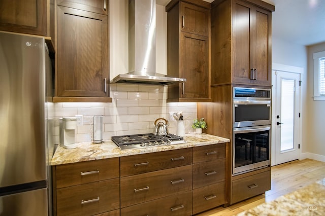 kitchen with wall chimney exhaust hood, stainless steel appliances, light stone countertops, and backsplash