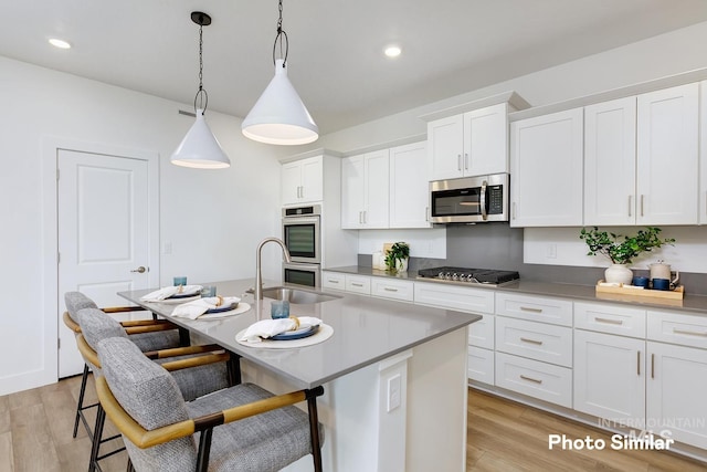 kitchen with white cabinets, a center island with sink, light hardwood / wood-style flooring, appliances with stainless steel finishes, and decorative light fixtures