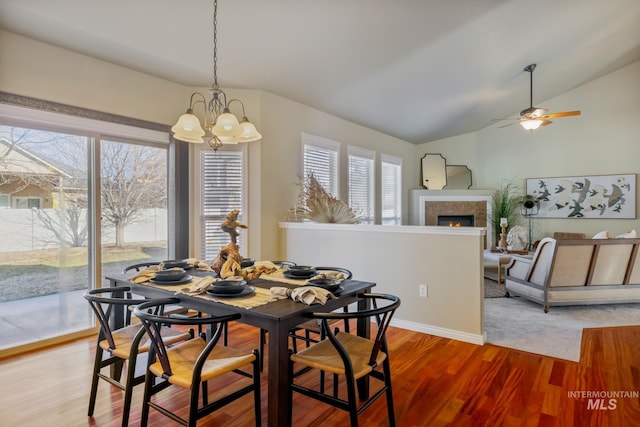 dining space featuring a tiled fireplace, hardwood / wood-style flooring, ceiling fan with notable chandelier, and vaulted ceiling