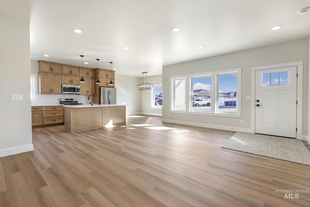 kitchen featuring appliances with stainless steel finishes, decorative light fixtures, tasteful backsplash, a kitchen island with sink, and light hardwood / wood-style flooring