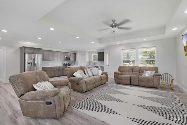 living room featuring a raised ceiling, a ceiling fan, recessed lighting, light wood-type flooring, and baseboards