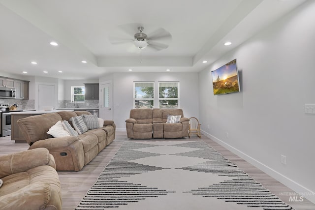 living area with recessed lighting, ceiling fan, baseboards, light wood-style floors, and a tray ceiling