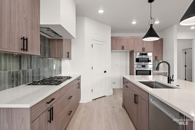 kitchen with wall chimney range hood, appliances with stainless steel finishes, a sink, light wood-style floors, and tasteful backsplash