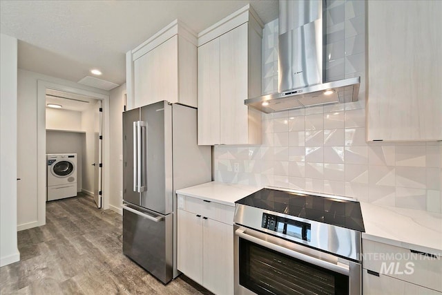 kitchen featuring light stone countertops, backsplash, stainless steel appliances, wall chimney range hood, and washer / dryer