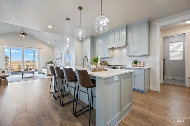 kitchen featuring vaulted ceiling, hardwood / wood-style floors, decorative light fixtures, backsplash, and a kitchen island with sink