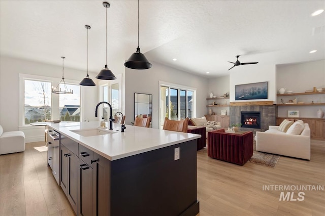 kitchen with sink, a tile fireplace, a kitchen island with sink, hanging light fixtures, and light wood-type flooring