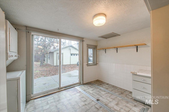 clothes washing area with a textured ceiling, stacked washer and dryer, and tile walls