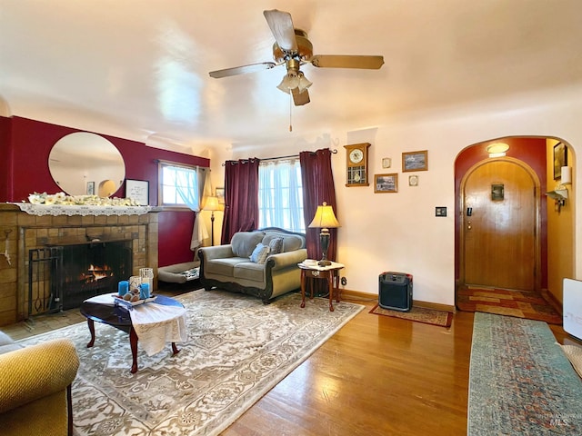 living room with a ceiling fan, a tiled fireplace, arched walkways, wood finished floors, and baseboards