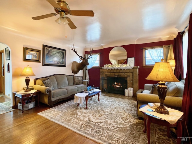 living room featuring arched walkways, ceiling fan, a warm lit fireplace, and wood finished floors