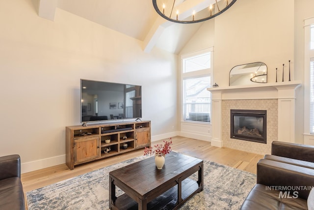 living room with high vaulted ceiling, a chandelier, a tiled fireplace, beam ceiling, and light hardwood / wood-style flooring