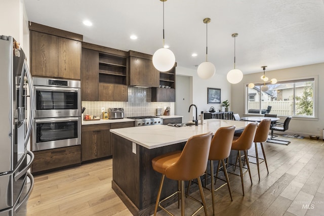 kitchen featuring light wood finished floors, open shelves, decorative backsplash, appliances with stainless steel finishes, and a kitchen bar