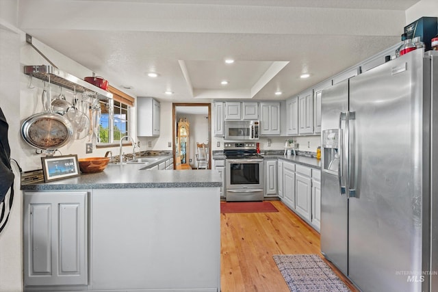 kitchen featuring appliances with stainless steel finishes, a sink, a peninsula, light wood-style floors, and a tray ceiling